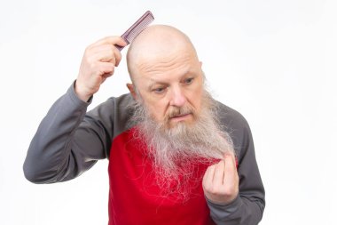 Uzun beyaz sakallı kel bir adam, kırmızı bir tarakla parlak kafa derisini tarıyor, kırmızı ve gri gömlekli, beyaz bir arka planda izole edilmiş, sakallı bir saç teli tutuyor..