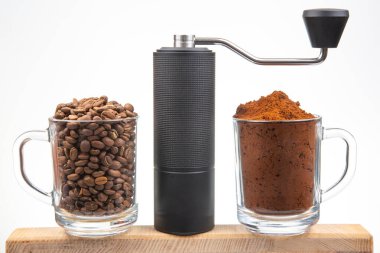 İki cam bardak, biri kahve çekirdekleriyle dolu, diğeri de öğütülmüş kahveyle, ahşap bir tahtanın üzerinde, kahve hazırlama sanatını gösteren ince bir manuel öğütücüye yan dön..