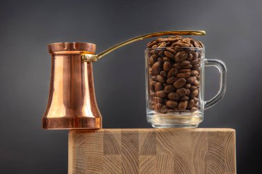 Geleneksel bir bakır çakmak ve kavrulmuş kahve çekirdekleri ile dolu bir cam bardak, koyu mavi arka planda, ahşap bir yüzeye oturur. Kırsal ve zarif bir kahve temalı kompozisyon..