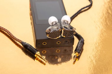 Siyah bir müzik cihazının üzerinde örülmüş kablo olan bir çift kulaklık, yansıtıcı altın arka plana ayarlı. Ses, teknoloji ve modern yaşam tarzı fotoğrafları için ideal..