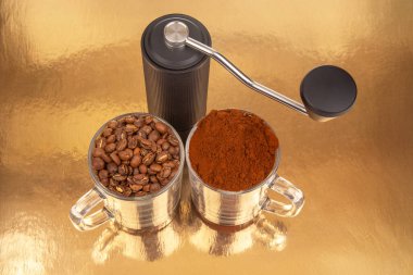 Bir el yapımı kahve öğütücüsü, biri kavrulmuş kahve çekirdekleri, diğeri taze öğütülmüş, altın bir yüzeyde, iki cam bardağın arasında oturur ve kahve öğütme işlemini gösterir.