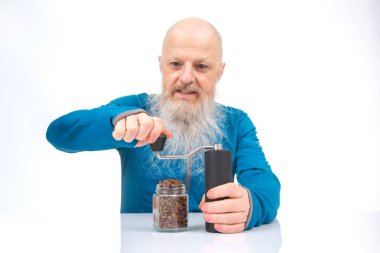 Uzun beyaz sakallı neşeli kel bir ihtiyar, gri kollu mavi bir gömlek giyerek kahveyi elle öğütür.