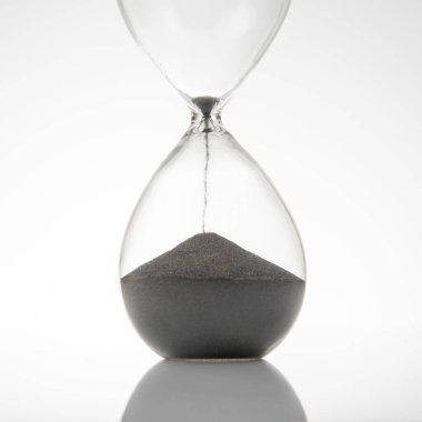 Kum saatine yakın çekim, koyu kumlar düşüyor, temiz beyaz arka plan üzerine kurulmuş. Zamanı, odağı ya da minimalizmi göstermek için harika.