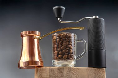 Bakır bir mağara, kahve çekirdekleri ile dolu bir cam bardak ve koyu arkaplana karşı ahşap bir blokta duran bir el yapımı kahve öğütücüsünün yer aldığı şık bir natürmort..