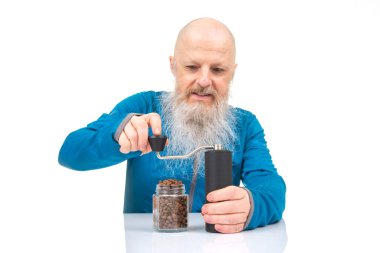 Uzun beyaz sakallı neşeli kel bir ihtiyar, gri kollu mavi bir gömlek giyerek kahveyi elle öğütür.