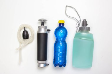 Beyaz arka planda siyah bir pompa, mavi plastik bir şişe ve kamp yapmak ve yürüyüş yapmak için ideal kayışlı yeşil bir hidrasyon şişesi de dahil olmak üzere bir dizi su arıtma aracı var..