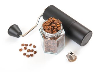 Metalik saplı siyah bir el değirmeni kavrulmuş kahve çekirdekleri, dağınık fasulyeler ve beyaz bir arka planda öğütücü ile dolu bir cam kavanozun yanında duruyor.