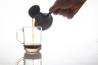 Bir el zarif bir şekilde geleneksel bir seramik çaydanlıktan temiz bir cam fincana zengin bir kahve döker, pürüzsüz beyaz bir arka plan ve ince bir yansıması vardır.