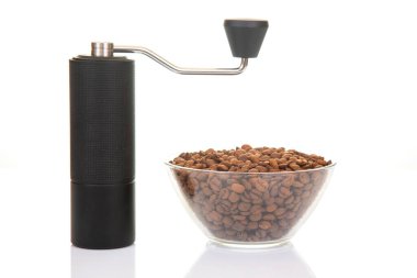 Süslü, siyah bir kahve öğütücüsü, cam bir kase taze kavrulmuş kahve çekirdeği ile eşleştirilmiş, bazıları etrafa dökülmüş, minimalist bir görünüm için temiz beyaz bir zemin üzerine kurulmuş.