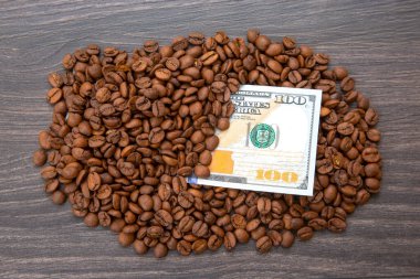 100 dolarlık banknot, koyu ahşap bir yüzey üzerinde kavrulmuş kahve çekirdekleri ile kısmen örtülmüş. Kahve ticareti, küresel ekonomi, kafein ticareti ve finansal yatırım kavramı