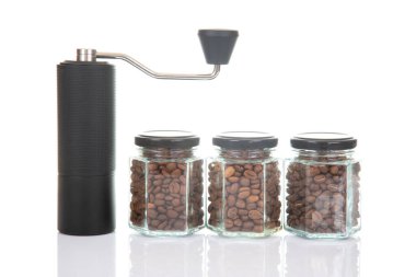 Kahve çekirdekleri için siyah manuel kahve öğütücüsü ve kavrulmuş kahve çekirdekleri ile dolu üç bardak şeffaf kavanoz ve beyaz arka planda kahve demlemek için toprak takımı..