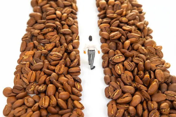 Kahve çekirdeği yığınları arasında yürüyen takım elbiseli minyatür bir figürün sembolik stok fotoğrafı kahve endüstrisindeki bir yolculuğu temsil ediyor. Beyaz arkaplan.