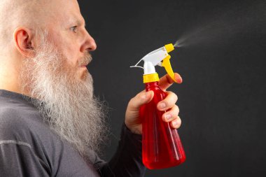 Kel ve sakallı bir adam koyu gri arka planda sprey şişesinden sıvı sıçratıyor.