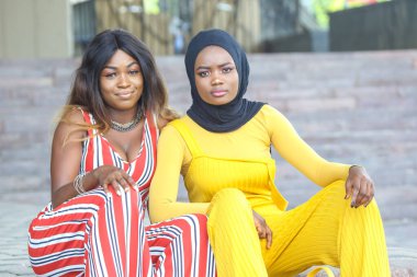 İki şık Afrikalı kadın yan yana oturmuş güneşli bir günün tadını çıkarırken temiz bir mimari geçmişe sahip çağdaş bir şehirde renkli kıyafetler sergiliyorlar..