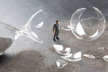 Takım elbiseli bir işadamının minicik bir figürü karanlık bir yüzeyde kırık cam parçalarının arasında, beklenmedik bir kaosun habercisi olarak duruyor. Çevredeki parçalar kazayı vurguluyor..