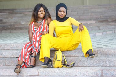 İki Afrikalı kadın taş basamaklarda rahatlar, canlı kıyafetler sergilerler. Biri tesettürlü sarı bir tulum giyerken, diğeri çizgili bir elbise giyiyor..