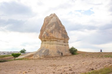 Türkiye 'nin Kapadokya kentinde bulutlu bir gökyüzüne karşı eşsiz bir kaya oluşumu ayakta duruyor. Geniş arazideki bu doğal harikanın ölçeğini gösteren iki birey yakınlarda yürüyor..