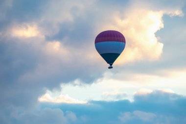 Canlı bir sıcak hava balonu, gün batımının yumuşak renkleriyle aydınlanan bulutlar arasında zarifçe sürüklenir..