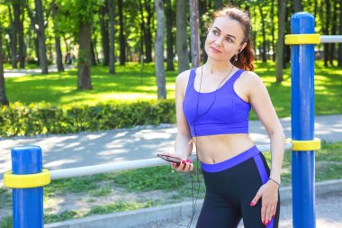 Spor kıyafetli genç bir kadın, canlı yeşil bir parkta açık hava egzersiz ekipmanlarına yaslanırken telefonuyla müzik dinlemekten hoşlanıyor. Ağaçlar ve yürüyüş yolu huzurlu bir ortam sağlar..