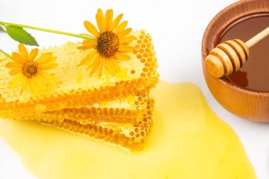 Taze bal peteği altın bal dolu ahşap bir kasenin yanında oturur, canlı sarı çiçeklerle süslenir, beyaz arka planda görsel olarak çekici ve doğal bir tatlılık gösterisi yaratır..