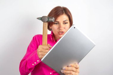Parlak pembe gömlek giyen bir kadın elinde çekiç tutarken ve tablete bakarken hayal kırıklığı gösterir. Stüdyo ayarı, onun ifadelerini vurgulayan düz beyaz bir arka plan içerir..