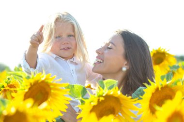 Bir ayçiçeği tarlasında, neşeli bir çocuk gülümseyen bir anne tarafından tutulurken gökyüzünü gösterir. Parlak güneş manzarayı aydınlatıyor, sıcak ve neşeli bir atmosfer yaratıyor..