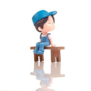 Mavi şapkalı ve tulumlu neşeli bir minyatür figür ahşap bir bankta oturuyor, parlak ve pürüzsüz bir ortamda huzurlu bir anın tadını çıkarıyor. Yansıtıcı yüzey olay yerine derinlik katıyor..