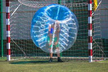 Büyük şeffaf balon kostümlü bir insan baloncuk futboluna katılmaya hazırlanıyor. Etkinlik, parlak güneş ışığı altında kale direkleriyle iyi korunan yeşil bir sahada gerçekleşiyor..
