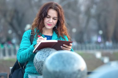 Kıvırcık saçlı, deniz mavisi ceket giymiş bir kadın, güneşli bir günde halka açık bir alanda pürüzsüz taş heykellerin yanında dururken bir deftere yazıyor..