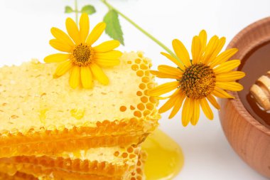 Damlayan ballı altın bal peteği canlı sarı çiçeklerin yanında sergilenir. Ahşap bir kase daha fazla bal içerir, mutfak için ideal taze ve doğal bir mutfak sahnesi yaratır..