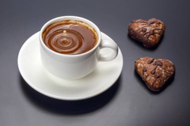 Bir fincan zengin sıcak çikolata, iki kalp şeklindeki kurabiyeyle birlikte beyaz bir fincan üzerinde oturur. Sıcak içecek dalgalanmalar yaratır, tatlı bir rahatlama anını davet eder..