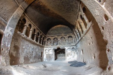 Ziyaretçiler Türkiye 'de antik bir kaya kesimi manastırı keşfederken, karmaşık kemerler ve oyulmuş taş duvarlar sergilenirken, güneş ışığı iç kesimin tarihi özelliklerini ve dokularını nazikçe aydınlatıyor..