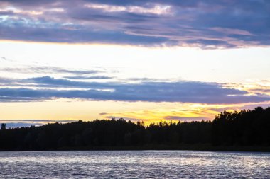 Güneş yemyeşil ağaçların arkasında batarken turuncu ve mor renkli canlı renkler gökyüzünü dolduruyor. Sakin su renkleri yansıtıyor ve akşam saatlerinde sakin bir atmosfer yaratıyor..