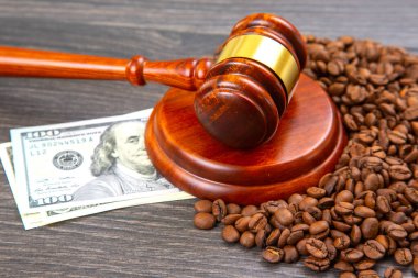 Ahşap bir tokmak, kavrulmuş kahve çekirdekleri ile çevrili dairesel bir zeminde duruyor. Yakınlarda 100 dolarlık banknot uzanıyor, karanlık bir ahşap yüzey üzerinde kanun ve ticaretin karşıtlığını oluşturuyor..