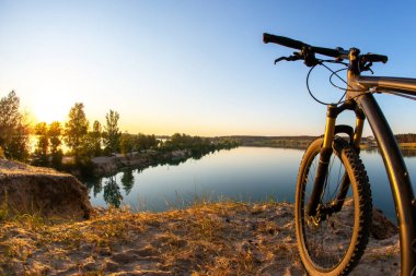 Güneş ağaçların arkasında batarken bir dağ bisikleti kumlu arazide dinleniyor. Sakin gölün üzerine sıcak renkler döküyor. Sakin su, akşam gökyüzünün canlı renklerini yansıtır..
