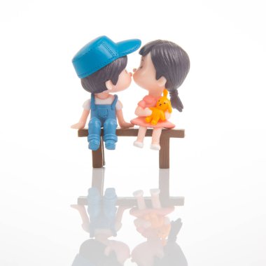 İki minyatür çocuk birbirine bakan küçük tahta bir bankta oturuyor. Hayal ürünü bir dünyayla çevrili şefkatli bir öpücüğü paylaşıyorlar, büyüleyici ve eğlenceli bir an yaratıyorlar..