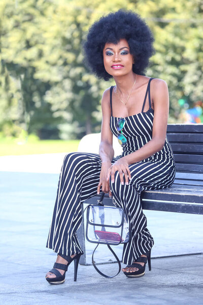 Уверенная в себе африканка сидит на скамейке в ярком городском парке, одетая в модный комбинезон в полоску и высокие каблуки.