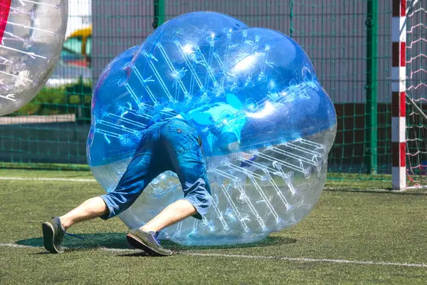 İki katılımcı yeşil bir çim sahasında balon futbolu oynuyorlar, parlak güneşin altında oyuncu bir rekabet ve eğlence sergiliyorlar. Birbirlerine çarptıklarında kahkahalar havayı doldurur..