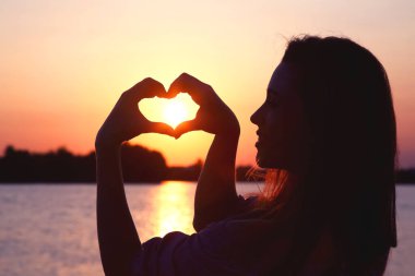 Bir kadın, elleri ile canlı bir günbatımına karşı bir kalp şekli yaratır, güzel bir siluet oluşturur. Sakin su, gökyüzünün sıcak renklerini yansıtıyor ve sakin bir atmosfer yaratıyor..