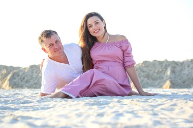 Gün batımında kumlu bir plajda dinlenen bir çift ebeveyn olmadan önce neşeli bir anı kucaklıyor. Kadın, bebek şişkinliğini şık bir elbiseyle gösterirken, erkek de yanında oturuyor..