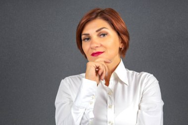 Beyaz gömlekli bir kadın, gri bir arka plana yaslanmış, çenesini elinin üzerinde dinlendiriyor..