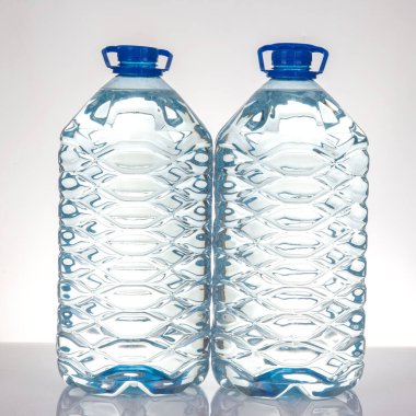 İki büyük, şeffaf su şişesi yan yana duruyor, pürüzsüz bir yüzeye ışığı yansıtıyor. Şişelerde desenli tasarımlar ve mavi başlıklar yer alıyor, tazelik ve su kaybını çağrıştırıyor..