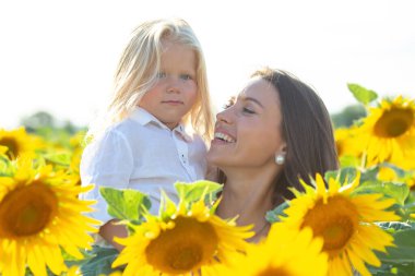 Sarışın çocuğunu açık gökyüzünün altında canlı bir ayçiçeği tarlasında tutan bir kadın. Anne sıcak bir şekilde gülümsüyor, ve çocuk meraklı bir şekilde bakıyor, doğada bir neşe ve bağlantı anı yaratıyor..