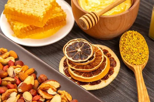 Canlı bir doğal bal gösterisi, kurutulmuş turunçgil dilimleri, çeşitli fındıklar, ve propolis koyu ahşap bir yüzeyde oturur, sağlıklı malzemelerin zengin renklerini ve dokularını sergiler..