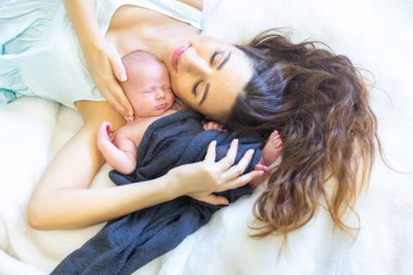 Bir anne yumuşak bir yatağa uzanıp yeni doğmuş bebeğini göğsüne yakın bir yere yatırır. Samimi an sıcaklığı ve sevgiyi yansıtır. Her ikisi de rahat ortamlarında huzur içinde dinlenir..