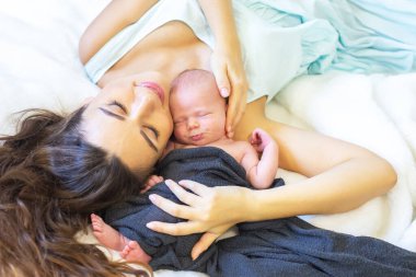 Yeni bir anne, yumuşak bir battaniyeye uzanmış, uyuyan bebeğini nazikçe tutarken gülümsüyor. Sıcak ve barışçıl atmosfer anne ve çocuk arasındaki bağın ve kapalı bir ortamın altını çiziyor..