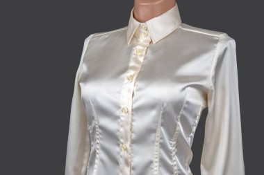 Klasik yakalı ve düğmeli bu zarif ipek bluz gri bir arka plana karşı bir mankenin üzerinde sergileniyor, pürüzsüz dokusu ve rafine tasarımı vurgulanıyor..