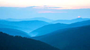 Yuvarlanan tepeler ve dağlar, güneş arka planda batarken yumuşak mavi renklerle yıkanır. Sisli manzaraya huzur ve yalnızlık atmosferi oluşturan sakin bir ışık yayar..