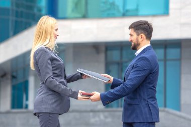 İki iş adamı, modern mimariyle çevrili profesyonel bir açık havada doküman alışverişinde bulunuyorlar. Gökyüzü açık ve aydınlık bir gün gibi görünüyor..