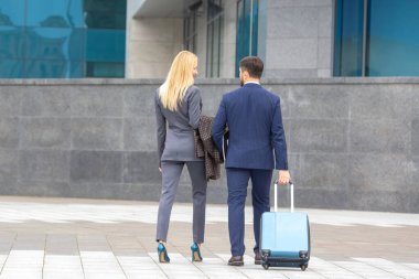 İş adamları bavullarını taşıyarak çağdaş bir yapıya doğru yürüyorlar. Erkek takım elbise giyip bavulu sürerken, kadın spor ayakkabıları ve spor ayakkabıları taşıyor..